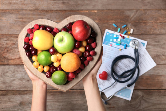 Female doctor holding plate with fresh fruits over wooden table, top view. Cardiac diet