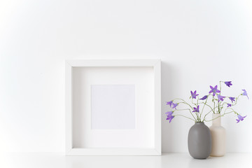 White square portrait frame mockup with spring bellflowers in vases near white wall on white background. Empty frame mock up for presentation design. Template framing for modern art.