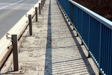 Bridge sidewalk with sand on cracked asphalt between guardrail on left and metal fence on right side on warm sunny day