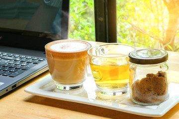 Hot cappuccino coffee cup, hot tea cup with brown sugar glass bottles and computer notebook on...
