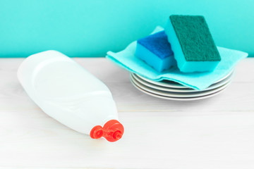 Bottle of dish washing, sponges, utensils on white wooden table against a green wall background,