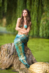 Gorgeous woman with long brown hair and dressed like a mermaid sits on the stone over water