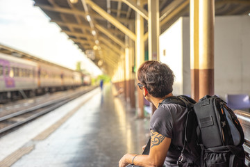 a traveller, backpacker man in casual clothes and sunglasses with a camera, sitting and waiting for a train at platform train station