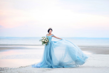 Stunning woman in rich blue dress stands on the wind on the sea shore