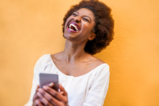 Young black woman standing isolated over yellow background using smart mobile phone - African girl laughing and smiling using web app on cellphone - Youth lifestyle and technology concept