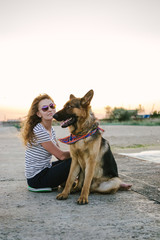 Happy young woman holding and sitting with her German shepherd dog outdoor on the sea pier during sunset