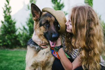 Happy young woman holding her funny German shepherd dog in hat, outdoor 