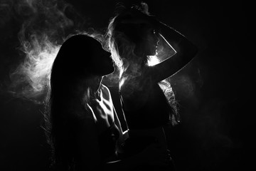 Silhouette of two sexy woman kissing holding in darkness through light and smoke