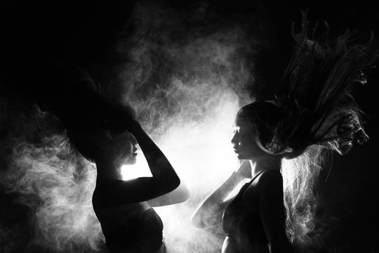 Silhouette of two sexy woman kissing holding in darkness through light and smoke
