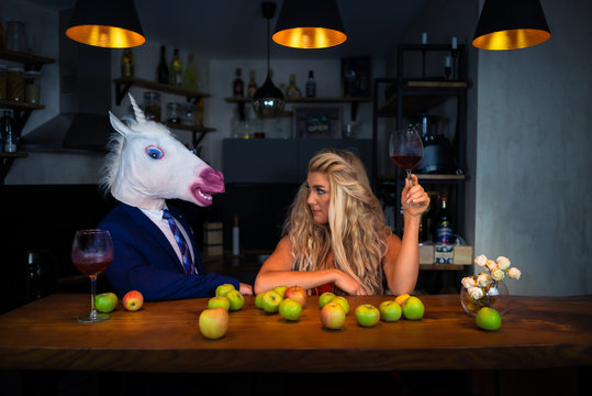 Unusual couple spend time together at the bar counter in stylish apartments with wine and food. Beautiful girl relaxing with funny boyfriend in comical mask. Unicorn in suit with young woman