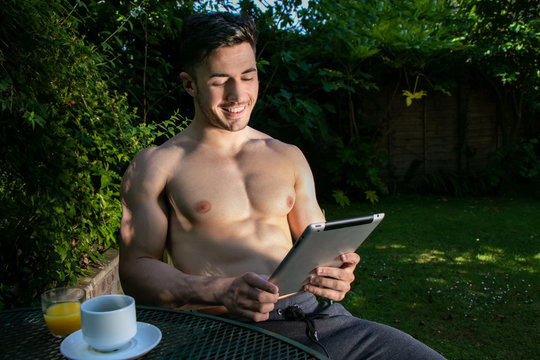 Hunky, Handsome Man With Pecs And Six Pack Abs Sits In Garden Drinking Coffee And Juice With Ipad Tablet