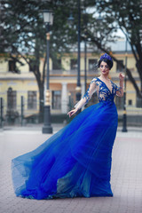 A beautiful sexy girl in a chic long blue dress developing in the wind on a city street.