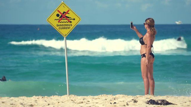 Bikini woman taking photo with smartphone on beach - danger sign and surfers