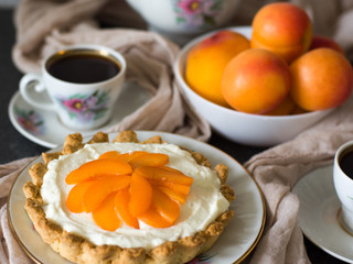 Cake with cream cheese with cream and apricots, against a background of mugs with coffee and apricots. Soft focus. Tart with apricots.