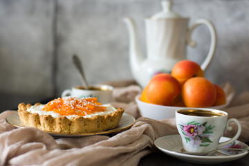 Cake with cream cheese with cream and apricots, against a background of mugs with coffee and apricots. Soft focus. Tart with apricots.