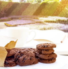Chocolate chip cookies and paper bag with cup coffee on table white with home garden background. With copy space for text.