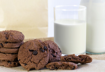 Chocolate chip cookies on brown paper and sack cloth with glass of milk and paper bag on white background.