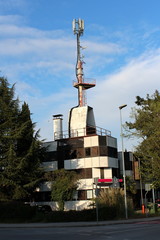 Cell phone tower mounted with old metal pole on top of building surrounded with trees and paved road