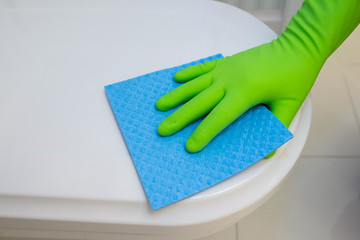 Woman in green rubber gloves cleaning toilet seat with blue cloth. Bathroom and toilet hygiene