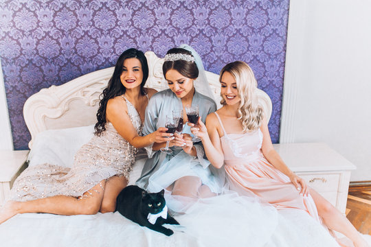 Happy wedding  bridesmaids and bride in silk robes sitting and lying on bed smiling and drink champagne. Celebrating wedding party at home before wedding ceremony. Black cat on bed.