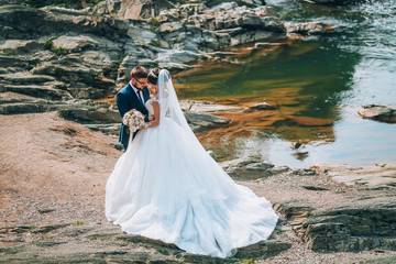 Romantic outdoor wedding shot of the fashionable newlywed couple hugging at the river bank. Bride...