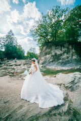 The bride in a wedding dress is standing on the mountain river bank with a bouquet of flowers....