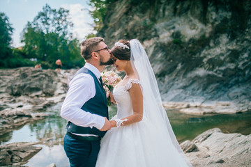 Romantic outdoor wedding shot of the fashionable newlywed couple hugging at the river bank. Bride...