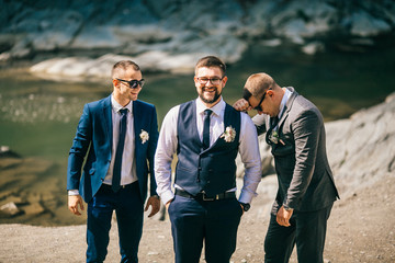 Groomsmen in black sunglasses hug groom tightly posing in the park. Funny wedding day moments of...