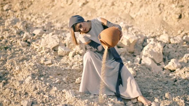 girl in ethnic dress sits against the background of stones near the road and pours sand out of the pitcher. The water turned into sand