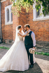 Beautiful newlyweds stand, embracing, near an old brick church with a large window. Portrait of a stylish groom and a young bride near an abandoned church.