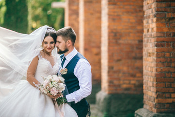 Bride and groom holding hands and walks near brick wall at wedding day. Newlyweds after wedding...