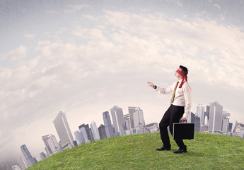 Young blindfolded businessman steps on a a patch of grass with a grey buildings in the background