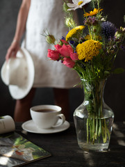 Bouquet of flowers in vase, cup of tea and magazine on wooden rustic table. Woman in white dress holding white hat on dark background.