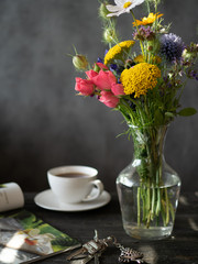 Bouquet of flowers in vase, cup of tea, magazine and bunch of keys on wooden rustic table.