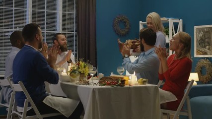 Cheerful woman bringing and serving huge roasted turkey on table having celebration of Thanksgiving with friends