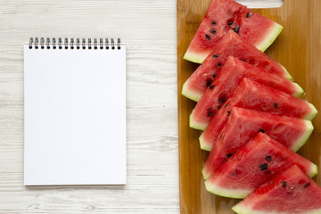 Sliced fresh ripe watermelon on cutting bamboo board with blank notebook over white wooden background, top view. From above.