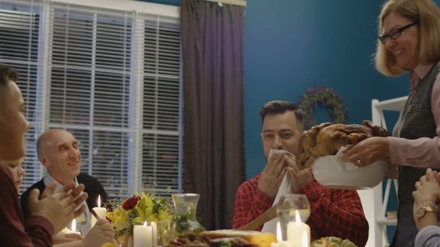 Mature woman serving plate with delicious roasted Thanksgiving turkey on table with cheerful family having traditional celebration