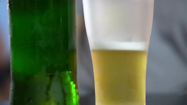 Frosted beer glass with golden drink inside. Green bottle close up 4K