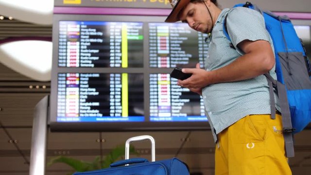 Young handsome international airport tourist passenger in hat uses mobile phone stands with backpack and suitcase at schedules of departure board of aircraft. 3840x2160