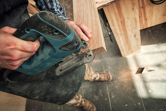 Close up of an experienced carpenter in work clothes and small buiness owner  is carving a wooden board on an  modern  hand drill in a light workshop side view, in the background a lot of tools