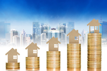 Wooden toy house putting on money coins stack arranged as a graph with blurred cityscape background; concept of money growth and saving money