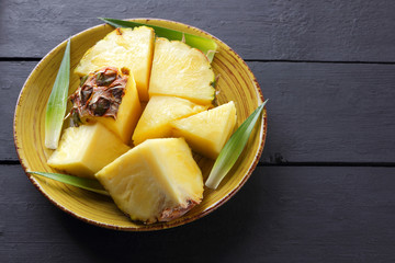 Pieces of pineapple on a yellow plate on wooden boards, pineapple fruit with green leaves on a dark background, tropical fruit salad for breakfast