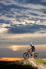 Side view of sporty cyclist sitting on mountain bike and observing beautiful landscapes and amazing sunset on cloudy sky. Man in black sportswear and helmet posing on rock hill.