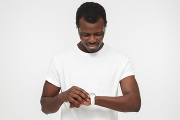 Young african american man wearing wireless headphones and blank white t shirt, checking smart watches with touch screen