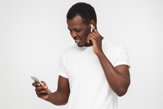 Portrait of handsome African American man in blank white t shirt, isolated on gray background, looking at screen of smartphone while listening to his favorite song