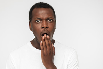 Close up photo of young african american man isolated on gray background dressed in blank white t shirt, covering mouth with hand, experiencing deep astonishment and fear
