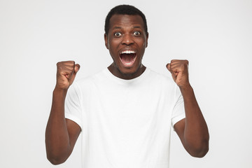 Emotional african american man in blank white t shirt isolated on gray background, clenching fists...