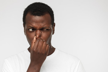 African american man in blank white tshirt, isolated on gray background holding his nose trying to escape from stinky smell and looking aside with worry looking for source of odor - obrazy, fototapety, plakaty