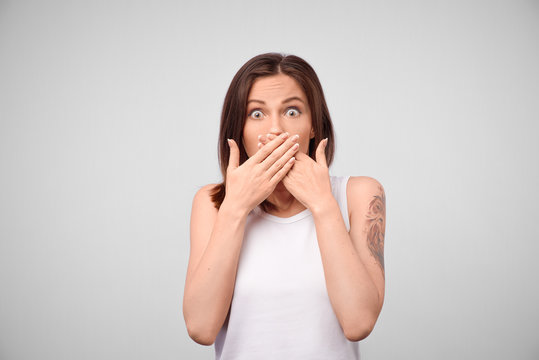 Portrait of a surprised young woman covering mouth with hands isolated over white background. Young woman covers mouth with hand, not tell, isolated on a gray background