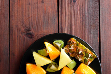 Fruit salad for vegans, pieces of pineapple, oranges and kiwi on a black plate, tropical fruits on wooden boards, citrus salad top view, vegetarian food